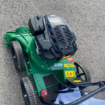 Top-down view of a green ride-on lawn mower on a gravel surface, showing the engine cover, control levers, and wheels for mowing. (Informative)