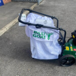Billy Goat lawn tool with a white protective cover over the handlebars in a paved parking area near a metal fence and brick wall to the left.