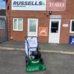 Brick building exterior with Russell's and Toro signs; a green KV Vacuum lawn vacuum on wheels parked in front near the reception door.