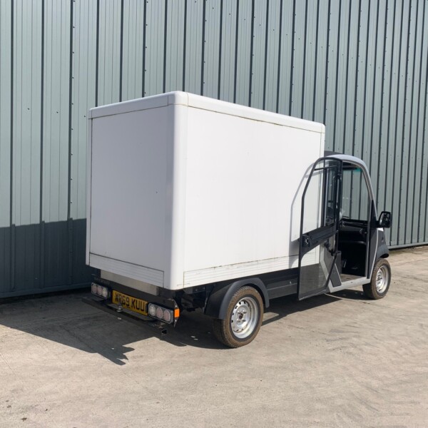 Small utility truck with a white box cargo area parked beside a tall gray corrugated-metal building.