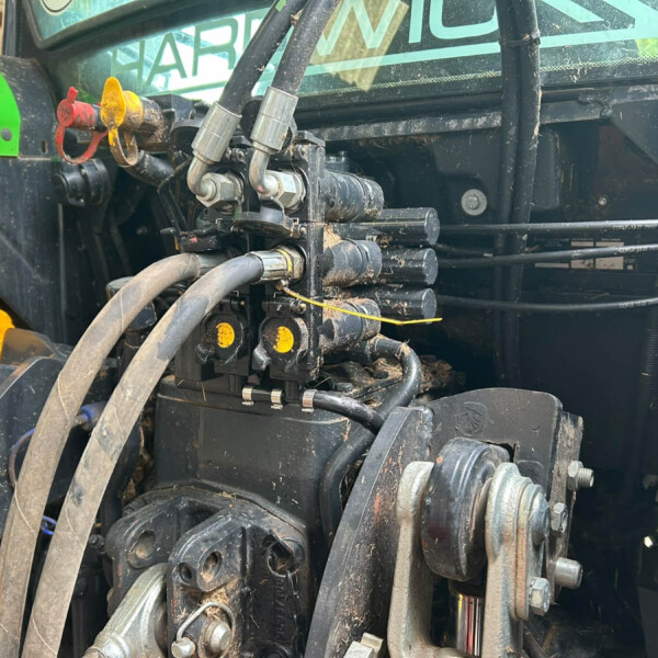 Close-up of hydraulic hoses, metal linkages, and control valves on a dusty green farm implement with safety sticker nearby.