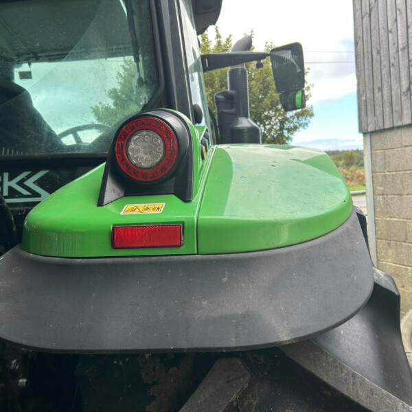 Close-up of a green tractor front end with a round red light, warning sticker, and exposed mechanical components near the hood edge.