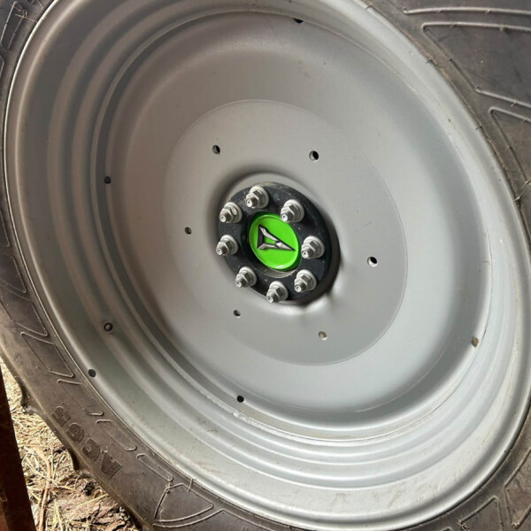 Close-up of a large tractor tire and gray wheel rim with a green center cap and eight lug nuts; a rusted metal beam is in the foreground on the left.