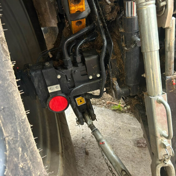 Underside view of a trailer hitch with hydraulic hoses, metal frame, and a red reflector under a muddy tire edge