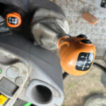 Close-up of an orange joystick control on heavy equipment, with numbered gear indicator (1–5) visible and blurred dashboard in the background.