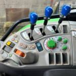Inside a tractor cab, three blue-topped gear levers rise from a gray control panel with gauges and colorful buttons.