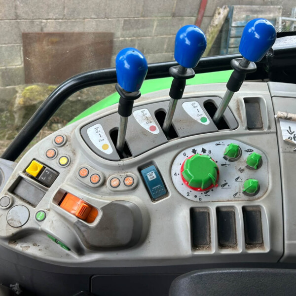 Inside a tractor cab, three blue-topped gear levers rise from a gray control panel with gauges and colorful buttons.