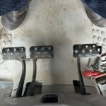 View of a vehicle's driver footwell with multiple pedals and scattered tools on a dirty metal floor