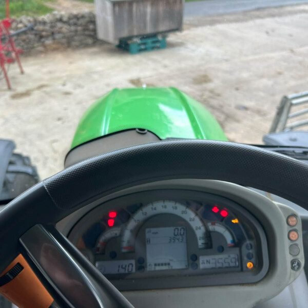 View from inside a tractor cab: green hood and dashboard with gauges, red farm equipment outside on a dirt surface.