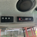Gray textured vehicle ceiling with a large circular black speaker, row of black switch panels to the left, and a Pioneer car stereo on the right; window and exterior structure visible through the side window.