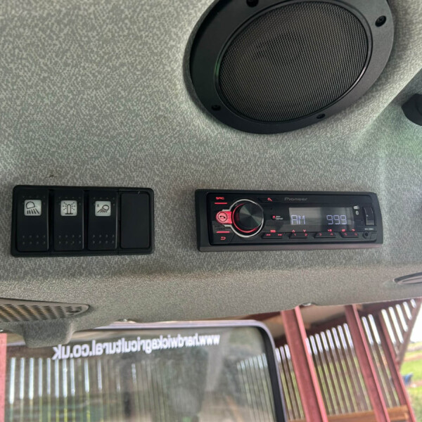 Gray textured vehicle ceiling with a large circular black speaker, row of black switch panels to the left, and a Pioneer car stereo on the right; window and exterior structure visible through the side window.
