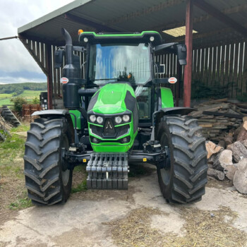 Front view of a large green tractor parked inside a shed, with stacked logs to the right.