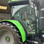 Close-up of a green agricultural tractor front with a large tire, cabin window, and metal exhaust stack in a shed.
