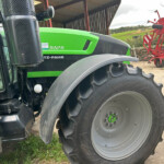 Close-up of a green tractor front with large rear tire and cab window, agricultural equipment in a shed in the background.