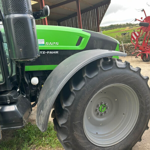 Close-up of a green tractor front with large rear tire and cab window, agricultural equipment in a shed in the background.