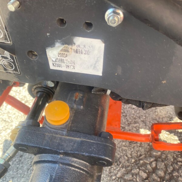 Close-up of a metal panel on a piece of equipment with a damaged sticker and bolts; an orange frame and a black hydraulic hose run nearby.