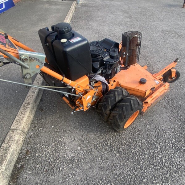 Orange tow-behind lawn mower attachment with a black gas engine, large rear tires, parked on asphalt near a metal gate.