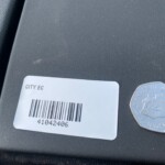 Close-up of a white barcode label on a dark textured surface; reads CITY EC with barcode 41042406, next to a silver octagonal token.