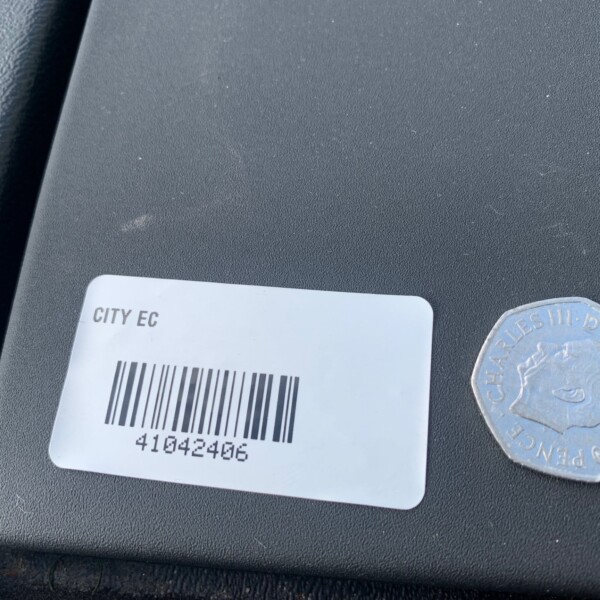 Close-up of a white barcode label on a dark textured surface; reads CITY EC with barcode 41042406, next to a silver octagonal token.
