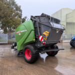 Green Fendt XtraCut farming header mounted on a trailer in a wet industrial yard with beige warehouse buildings in the background.