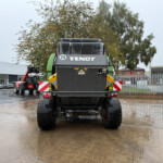 Back view of a large green and gray Fendt agricultural machine in a wet yard, with brick fences, striped hazard panels, and trees in the background.