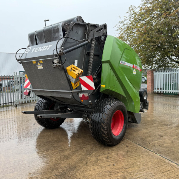 Rear view of a green Fendt agricultural harvester header with red wheels on a wet concrete lot behind a metal fence.