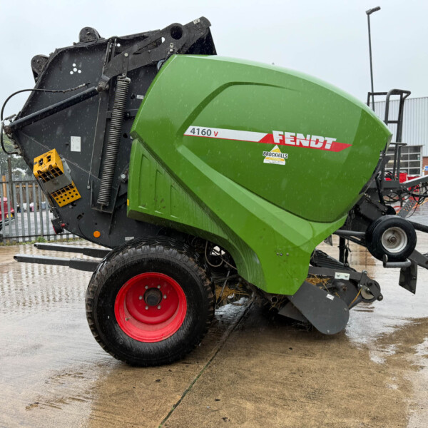 Green Fendt 4160 V combine header lying on its side in a parking lot, red wheels, and rain falling. The machine's dark frame and yellow steps are visible in the foreground.