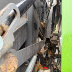 Close-up view inside industrial equipment showing belts, hoses, and metal components with wood debris and a bright green panel on the right side.