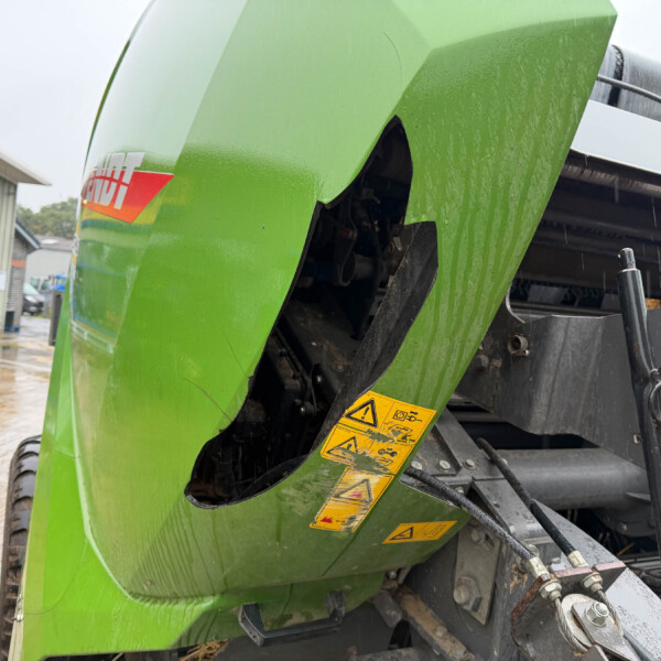 Close-up of a green combine harvester body with yellow warning stickers and exposed machinery in a rainy outdoor lot.