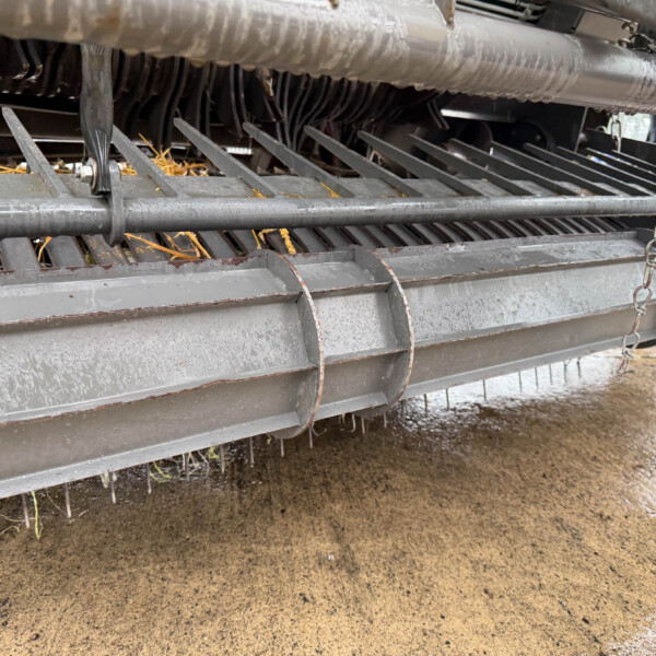 Close-up of the underside of a grain harvester with metal teeth and a chain, showing dirt and straw debris gathered between the blades.