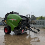 Green Fendt combine harvester parked on a wet concrete yard with a metal fence in the background and an overcast sky, rain droplets visible on the equipment.