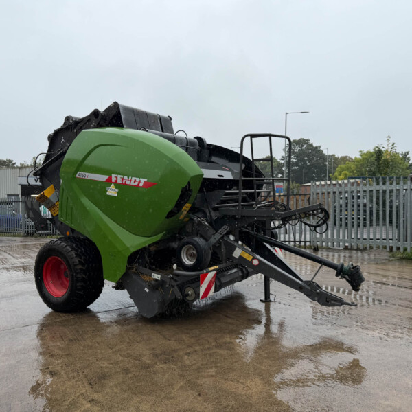 Green Fendt combine harvester parked on a wet concrete yard with a metal fence in the background and an overcast sky, rain droplets visible on the equipment.