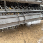 Close-up of a gray agricultural header with vertical metal tines resting above a wet, muddy ground. It shows a chain and protective bars.
