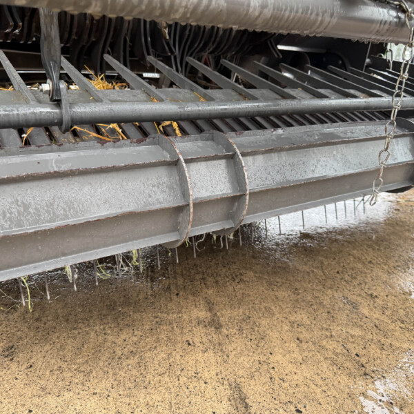 Close-up of a gray agricultural header with vertical metal tines resting above a wet, muddy ground. It shows a chain and protective bars.