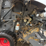 Close-up of a large agricultural machine's inner mechanisms, with belts, pulleys, hoses, and a bright red wheel, with straw around the base.