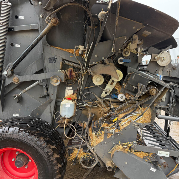 Close-up of a large agricultural machine's inner mechanisms, with belts, pulleys, hoses, and a bright red wheel, with straw around the base.