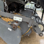 Close-up of a gray farming machine's cutting/feeding mechanism with chains, bolts, and straw debris on the metal frame.