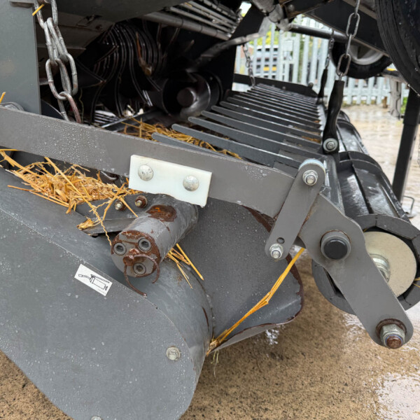 Close-up of a gray farming machine's cutting/feeding mechanism with chains, bolts, and straw debris on the metal frame.