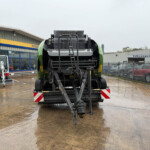 Front view of a large green agricultural harvester on a wet showroom lot, with New Holland Agriculture signage and a Volkswagen building in the background.