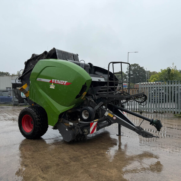 Green Fendt combine harvester parked in a fenced industrial yard on a rainy day, wet ground visible behind it.