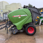 Green Fendt 4160V baler on a trailer in an industrial yard with a New Holland Agriculture sign in the background.
