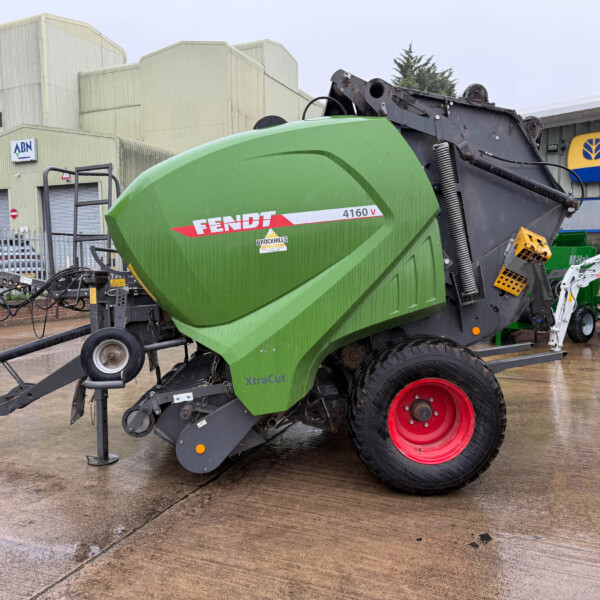 Green Fendt 4160V baler on a trailer in an industrial yard with a New Holland Agriculture sign in the background.