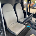 Interior of a bus cockpit with two gray driver seats, steering wheel, and ignition keys visible in the foreground outside the door window.