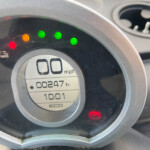 Close-up of a car instrument cluster: speedometer at 0 mph with green indicator lights and a red parking brake warning.