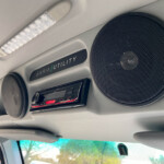 Inside a car ceiling showing a large round speaker, an overhead white textured light, and a panel labeled 'GARIA UTILITY'.