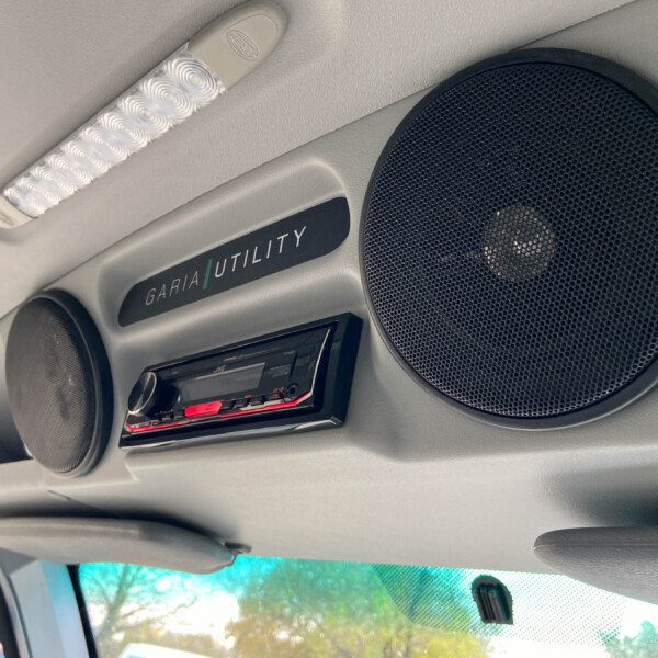 Inside a car ceiling showing a large round speaker, an overhead white textured light, and a panel labeled 'GARIA UTILITY'.