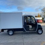 White delivery box truck with the cab door open, parked in a lot with other vans and cars nearby.