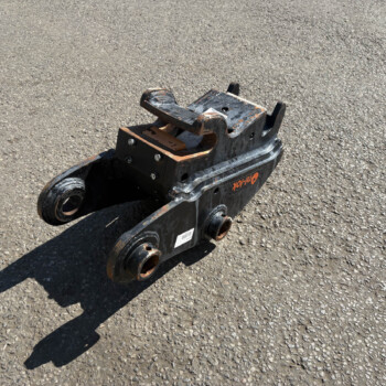 Hydraulic breaker attachment resting on rough asphalt surface, angled view shows worn metal and bolts.