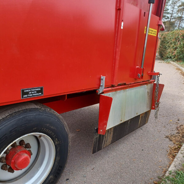 Close-up of a red trailer showing a large wheel, chain and latch hardware along the side, with hedge and road in the background