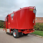 Red tanker trailer parked on a street, with a hedge and tiled-roof building in the background.
